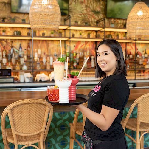 A person is holding a tray with colorful drinks in a vibrant bar with wicker chairs, decorative lights, and a patterned bar counter, smiling at the camera.