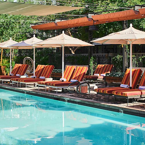 The image shows a swimming pool with lounge chairs and umbrellas lined up along the poolside, surrounded by greenery and outdoor decor.