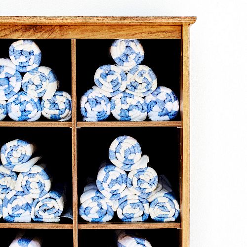 The image shows a wooden shelf with several compartments, each containing neatly rolled blue and white towels stacked in a pyramid shape.