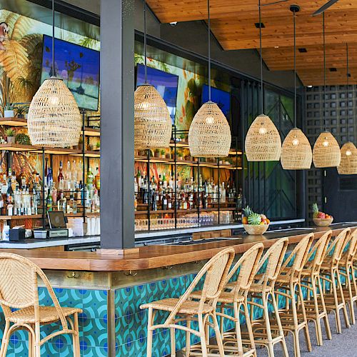 The image shows a stylish bar with rattan chairs, hanging lamps, and a stocked bar counter. The ceiling has exposed wood and fans.