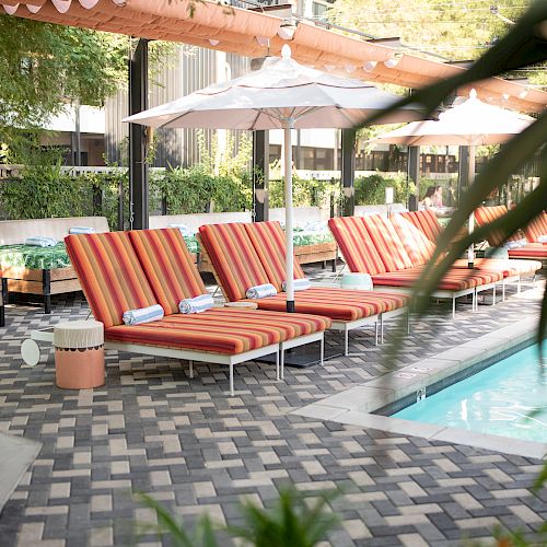 The image shows a poolside area with orange striped lounge chairs, white umbrellas, and a checkered tile floor, surrounded by greenery and buildings.