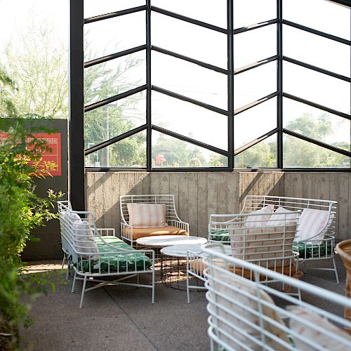 A modern, sunlit indoor seating area features wire-frame chairs with cushions, small wooden tables, large windows, and ceiling fans.