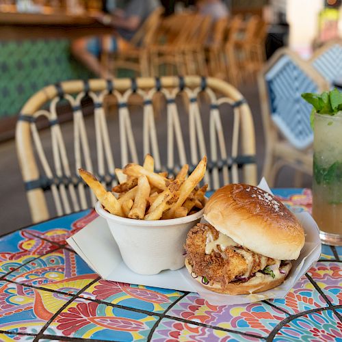 A table with a colorful pattern has a serving of fries, a burger, and a drink with mint leaves beside an empty chair in a cozy setting.