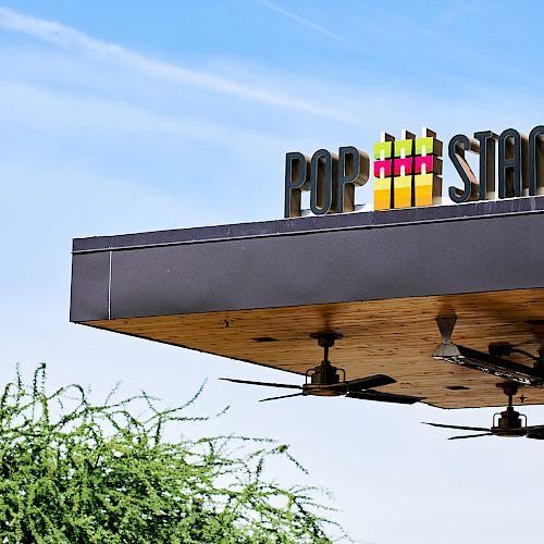 The image shows a building's edge with a sign reading "POP STAND!" under a blue sky with some clouds and a tree visible below.