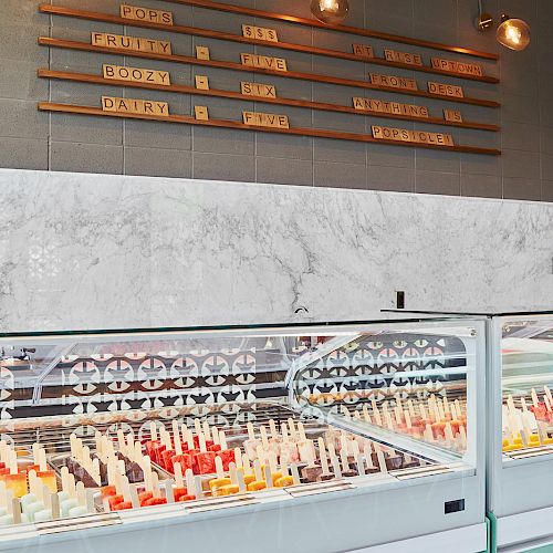 This image shows an ice cream or popsicle display case with a marble counter and a menu board above it, inside a brightly lit shop.