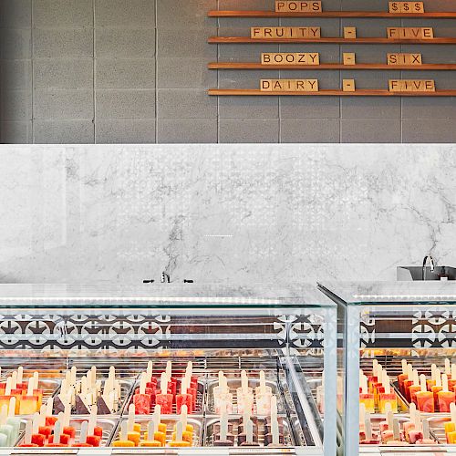 The image shows a modern ice cream shop interior with glass display freezers filled with colorful popsicles and ice cream, and a menu board on the wall.