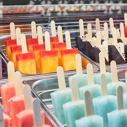 The image shows a display of colorful popsicles, neatly arranged in metal trays, with various flavors including blue, purple, red, and yellow.