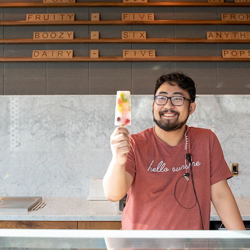 A smiling person behind a counter, holding up a colorful popsicle, with a menu board in the background displaying various options.