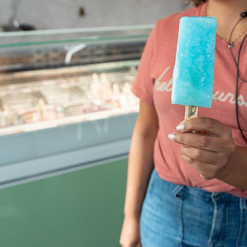 A person is holding a blue popsicle in front of a display case at what appears to be an ice cream shop, wearing a peach-colored shirt and jeans.