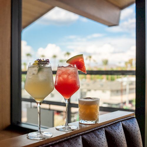 The image shows three distinct cocktails on a table by a window, offering a view of rooftops and palm trees under a clear blue sky.