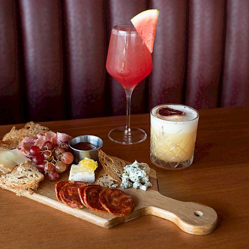 A charcuterie board with meats, cheeses, and bread is accompanied by a pink cocktail with a watermelon garnish and a creamy cocktail.