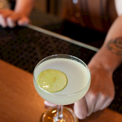 A person is holding a cocktail glass with a lime slice garnish, standing at a bar counter. The background shows blurred details of the bar setting.