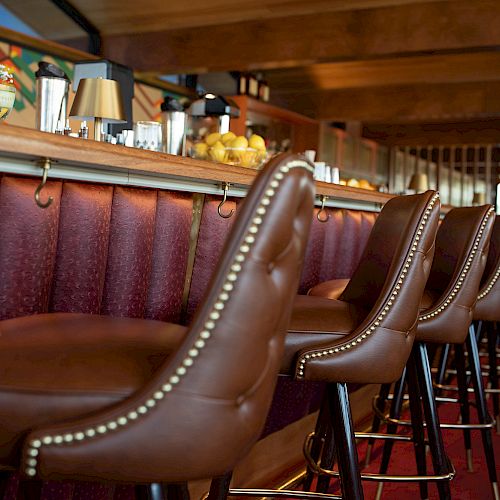 The image shows a row of brown leather bar stools with nailhead trim at a restaurant bar, with various bar items and fruit visible on the counter.