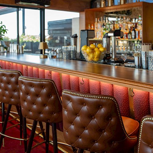 A bar with cushioned stools, a long counter, bottles of alcohol, glasses, and a bowl of lemons, and windows in the background.