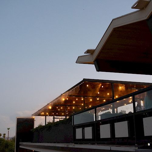 An outdoor terrace with string lights hanging above, located on a modern building with a clear evening sky in the background.