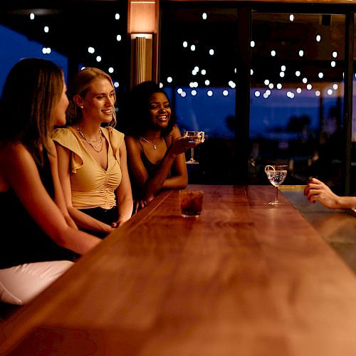 Three women are sitting at a bar, chatting and smiling, while the bartender serves them drinks in a warmly lit atmosphere.