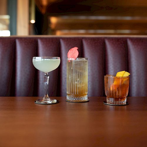 Three cocktails are lined up on a wooden table, with a cushioned booth in the background.