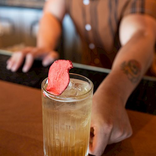 A person serving a cocktail garnished with a pink item in a glass at a bar. The focus is on the drink, with the person in the background blurred.