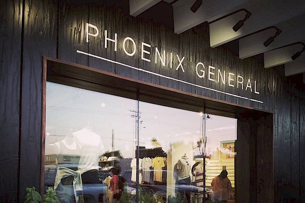 The image shows the storefront of a shop named "Phoenix General", featuring a display window with clothing and potted plants inside.