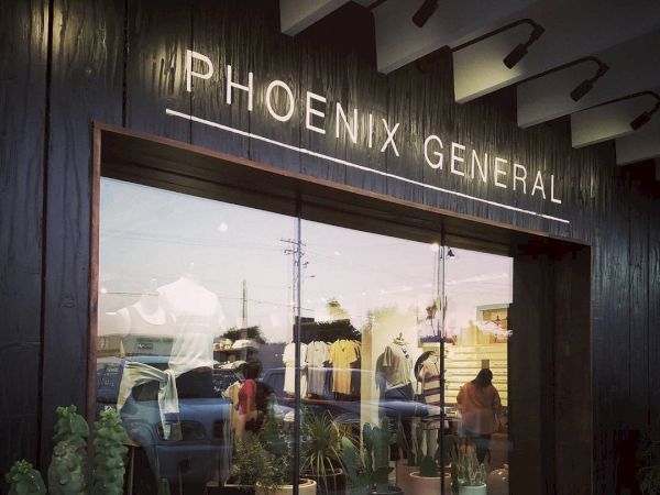The image shows the storefront of a shop named "Phoenix General", featuring a display window with clothing and potted plants inside.