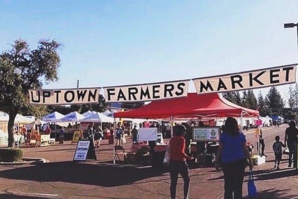 People are walking under a banner that reads "Uptown Farmers Market" with various vendor tents set up in the background.