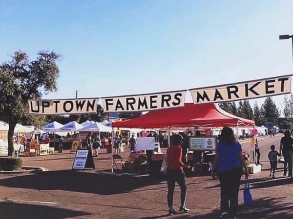 People are walking under a banner that reads "Uptown Farmers Market" with various vendor tents set up in the background.
