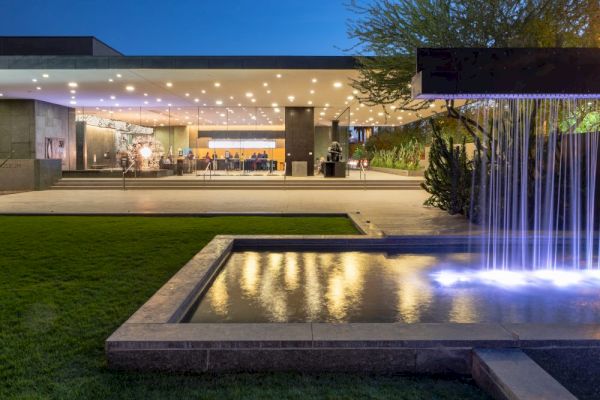 A modern building entrance with a well-lit exterior includes a reflecting pool with a waterfall feature and lush green surroundings.