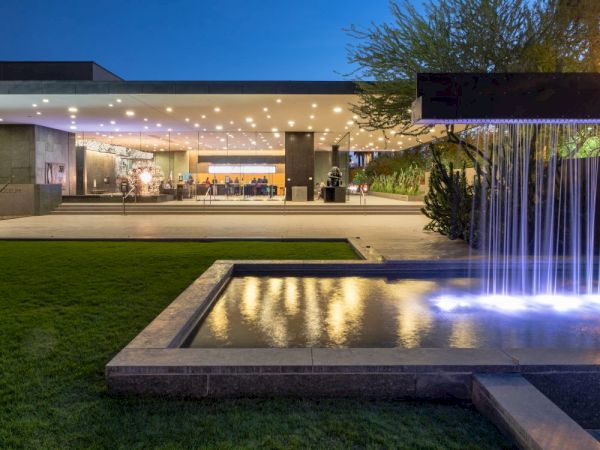 A modern building entrance with a well-lit exterior includes a reflecting pool with a waterfall feature and lush green surroundings.