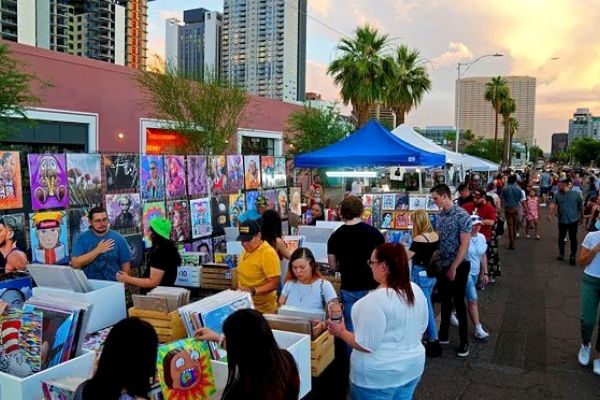 A vibrant outdoor art market with people browsing through paintings. Buildings and palm trees are visible in the background under a colorful sky.