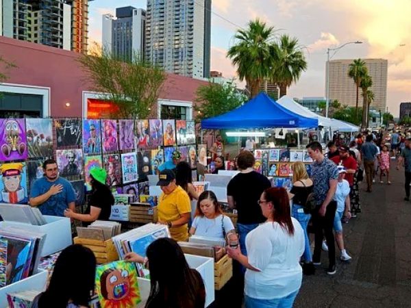 A vibrant outdoor art market with people browsing through paintings. Buildings and palm trees are visible in the background under a colorful sky.