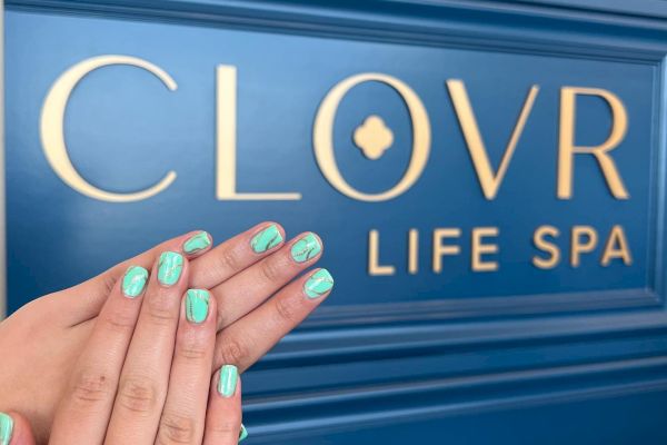 A pair of hands with aqua manicured nails is shown in front of a blue wall with the sign "CLOVR LIFE SPA" in gold lettering.