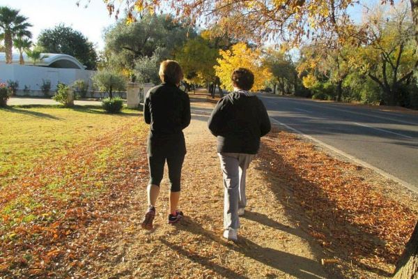 Two people are walking on a path surrounded by autumn foliage, with sunlight casting long shadows in the late afternoon.