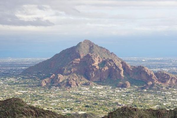 The image shows a rocky mountain in the backdrop with a cityscape at its base, surrounded by smaller hills and a cloudy sky.