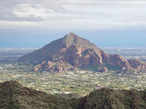 The image shows a rocky mountain in the backdrop with a cityscape at its base, surrounded by smaller hills and a cloudy sky.
