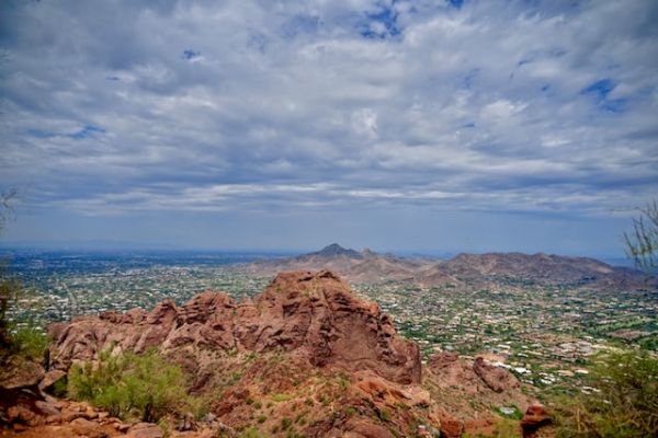 A scenic view of a rocky landscape with scattered greenery, under a partly cloudy sky. In the distance, a city and more mountains are visible.