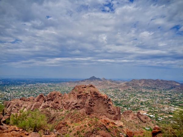 A scenic view of a rocky landscape with scattered greenery, under a partly cloudy sky. In the distance, a city and more mountains are visible.