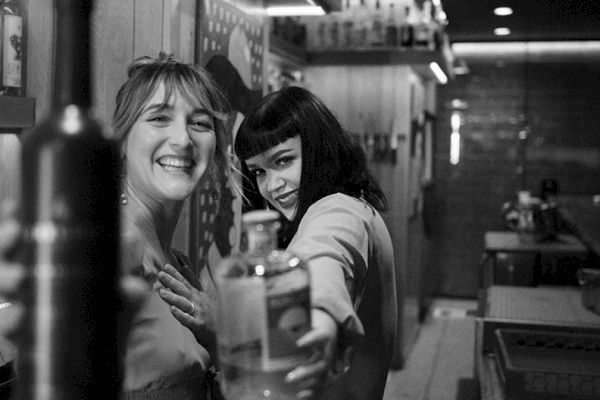 Two women are smiling and posing in a bar, one holding a bottle towards the camera. The setting appears to be a modern, cozy bar.