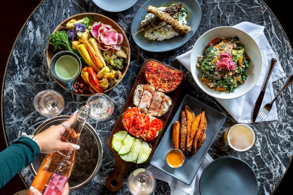 A table set with assorted dishes including salads, grilled vegetables, bread, sauces, sweet potato fries, and a person pouring a drink.