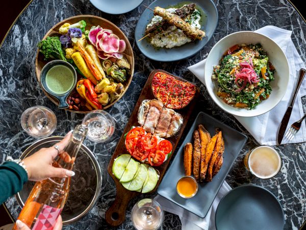 A table set with assorted dishes including salads, grilled vegetables, bread, sauces, sweet potato fries, and a person pouring a drink.