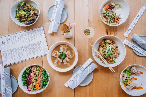 An overhead view of a dining table with various dishes including salads, noodles, and dumplings, accompanied by chopsticks and a menu on a wooden surface.