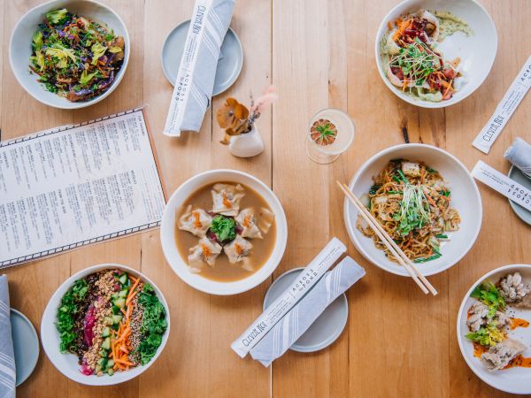 An overhead view of a dining table with various dishes including salads, noodles, and dumplings, accompanied by chopsticks and a menu on a wooden surface.