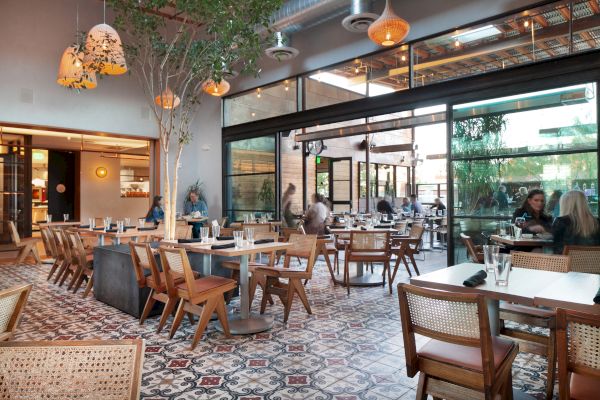 A modern restaurant with wooden tables and chairs, pendant lighting, tiled floors, and large windows. Some customers are seated and enjoying their meals.