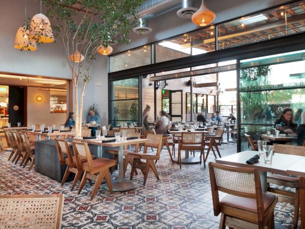 A modern restaurant with wooden tables and chairs, pendant lighting, tiled floors, and large windows. Some customers are seated and enjoying their meals.