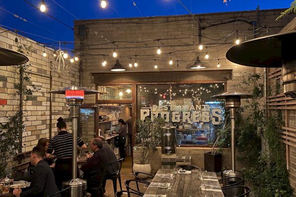 A cozy outdoor dining area with string lights, people sitting at tables, and a rustic brick wall backdrop, offering a warm and inviting atmosphere.