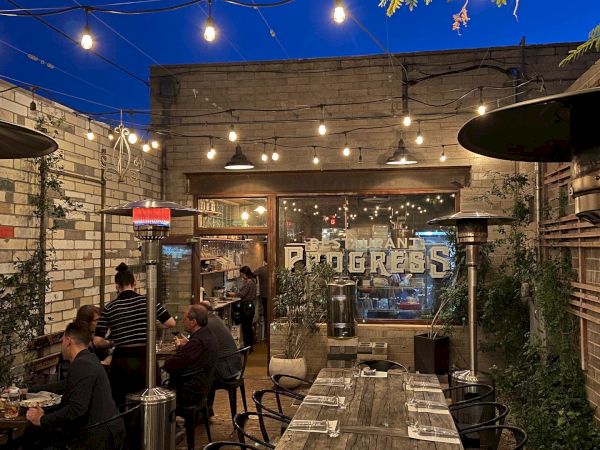 A cozy outdoor dining area with string lights, people sitting at tables, and a rustic brick wall backdrop, offering a warm and inviting atmosphere.