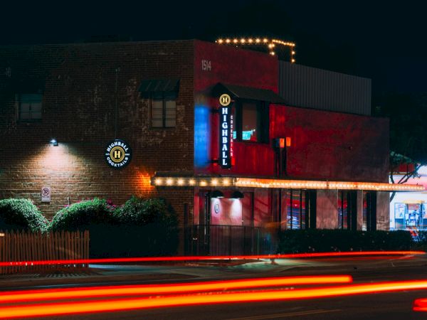 Nighttime photo of a brick building with neon signs reading "Highball," and light trails from passing cars, exuding a vibrant nightlife scene.