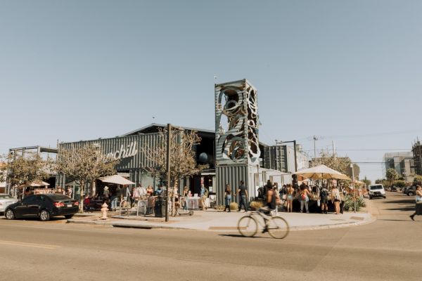 An outdoor scene of a busy modern building with a distinctive tower, people gathering, umbrellas, parked cars, and a bicyclist passing by.