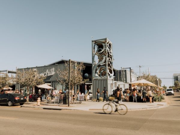 An outdoor scene of a busy modern building with a distinctive tower, people gathering, umbrellas, parked cars, and a bicyclist passing by.