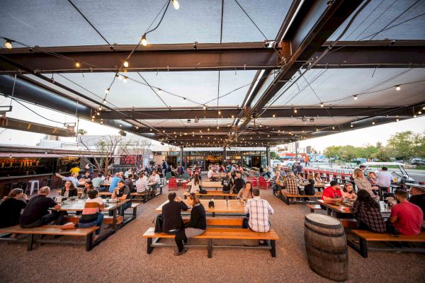 A bustling outdoor seating area under string lights, filled with people at wooden tables and benches, with food trucks and greenery in the background.