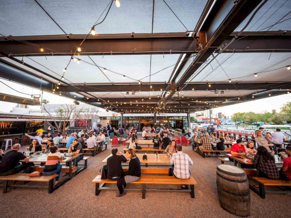A bustling outdoor seating area under string lights, filled with people at wooden tables and benches, with food trucks and greenery in the background.